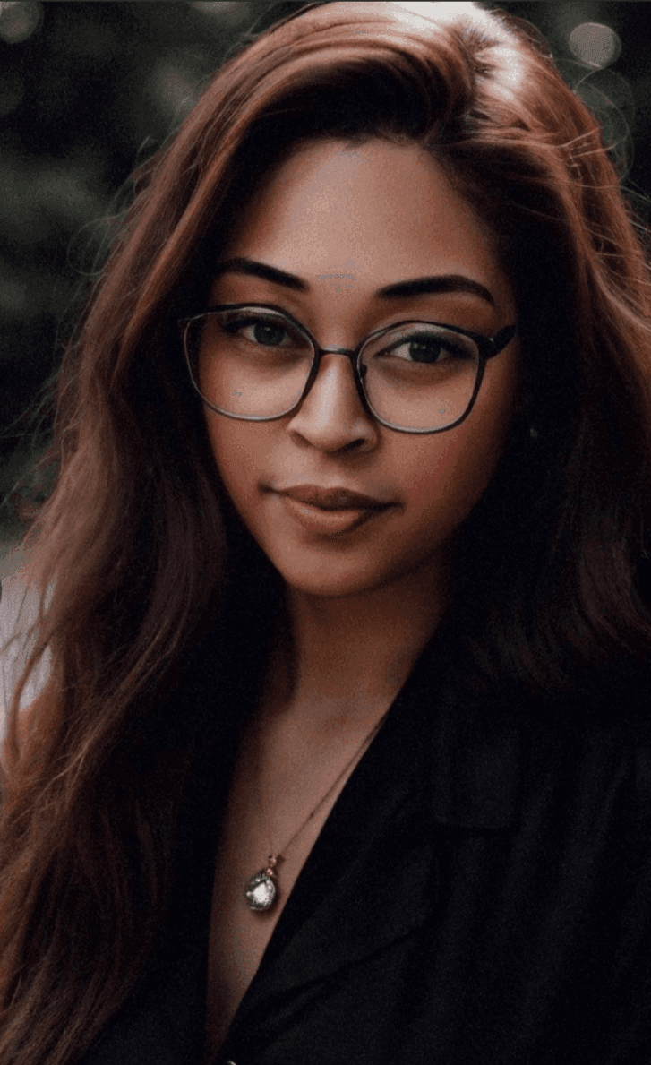 Woman with glasses and long hair, wearing a pendant necklace, against a blurred background.